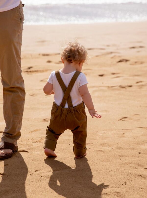 Anonymous dad looking after small kid carefully stepping on sandy beach in sunny day